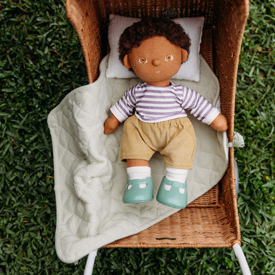 Dinkum Dolls doll resting on a sage green quilted blanket with a white pillow in a woven strolley outdoors on grass