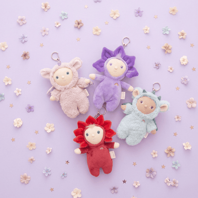 Four Dinky Dinkums plush bag charms in pink purple blue and red flower inspired outfits arranged on a lavender background with small floral decorations