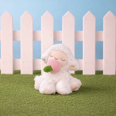 Binky Pets white lamb plush toy with closed eyes and a pink flower in its mouth sitting on green grass in front of a pastel pink picket fence