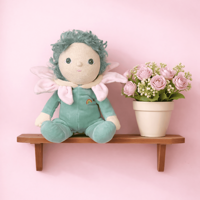 Dinky Dinkums Lily doll in a pale green outfit with pink wings sitting on a wooden wall shelf beside a pot of pink flowers against a soft pink background