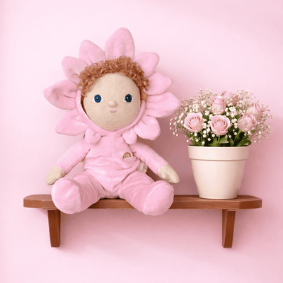 Dinky Dinkums Magnolia flower doll in a pink floral outfit seated on a wooden wall shelf beside a pot of delicate pink flowers against a soft pink backdrop