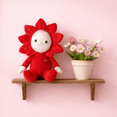 Dinky Dinkums Rose doll in a red flower outfit sitting on a wooden shelf beside a potted bouquet of pink and white flowers against a soft pink wall