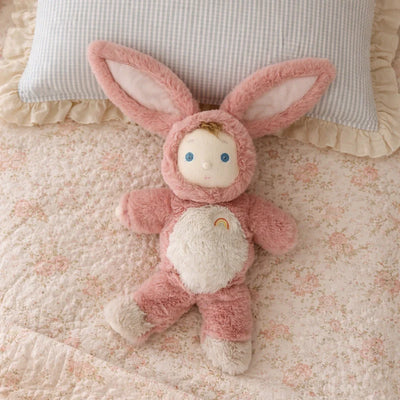 Pink bunny soft toy with floppy ears and blue eyes lying on a floral bedspread beneath a checked pillow from Cozy Day Dinkums