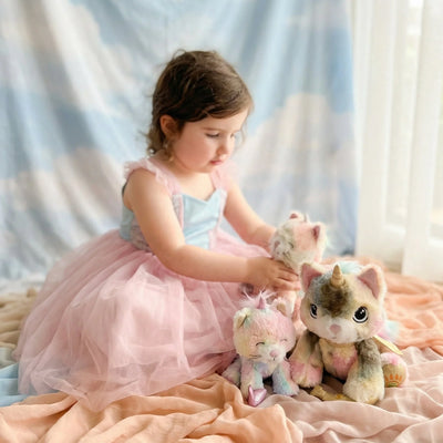 Child in a pink dress cuddling three soft kittycorn plush toys including Sprinkle in a cozy bedroom setting from Dinkum Pets for imaginative play