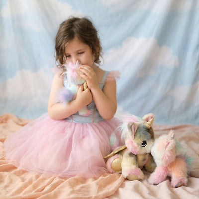 Child in a pink tulle dress cuddling a rainbow plush caticorn beside two pastel caticorn toys on a bed for imaginative play by Dinkum Pets