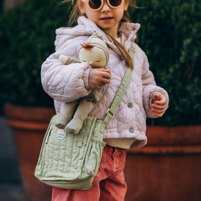 Child wearing sunglasses carries a soft doll and sage green quilted changing bag across the body outdoors from Dinkum Dolls