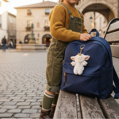 Child beside a navy backpack with a soft white lamb bag charm clipped to the front pocket on a wooden bench in a cobbled town square
