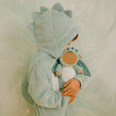 Child wearing a soft mint dragon costume cuddles a small plush dragon toy with shimmery wings. Cozy Dinkums imaginative play scene.