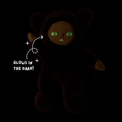 A dark plush teddy bear with glowing green eyes and a small stitched smile, shown on a black background with the text 'GLOWS IN THE DARK!' and sparkles pointing to its eyes.