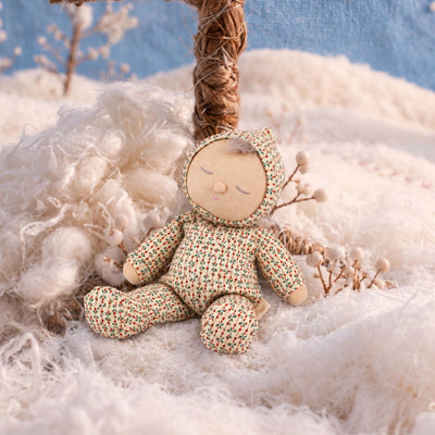 Soft baby doll with sleepy embroidered closed eyes, a smiling mouth and a tuft of brown hair, wearing a cream footed sleepsuit and matching bonnet printed with small red and green sprigs.
