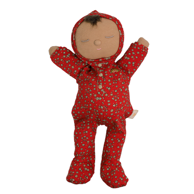 Soft baby doll with sleepy embroidered closed eyes, a smiling mouth and a tuft of dark brown hair, wearing a red footed sleepsuit and matching bonnet with a ditsy floral print, button placket, a small embroidered rainbow, and arms raised.