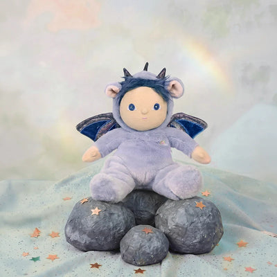Dinky Dinkums Dusty Dragon plush doll in a lavender dragon costume sitting on grey rocks with wings and stars in a soft pastel Rainbow Realm setting