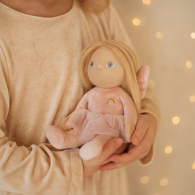 Dinky Dinkums Fairy Fiona doll with blonde hair and embroidered rainbow held in a child’s arms wearing a sparkly pink onesie and soft tulle skirt