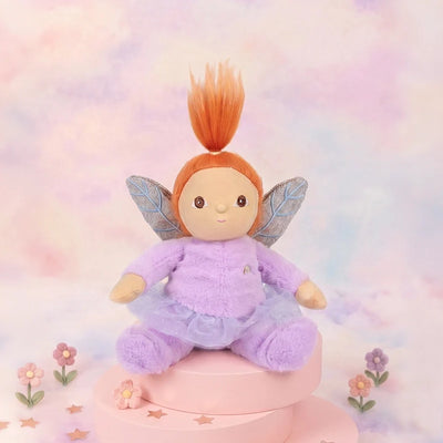 Dinky Dinkums Flora Fairy plush doll with orange hair lavender outfit and delicate wings seated on a pink pedestal surrounded by pastel flowers