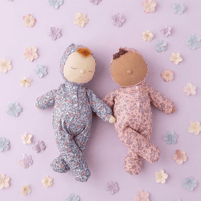 Two Dozy Dinkums soft dolls in blue and blush floral sleepsuits lying side by side on a pastel pink background with scattered fabric flowers
