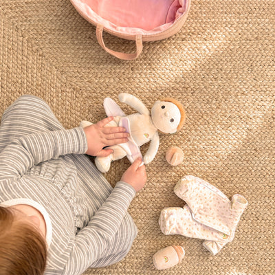 A child dresses a small Binky Dinky doll on a woven mat with spare clothes nearby and a pink carry bag above them