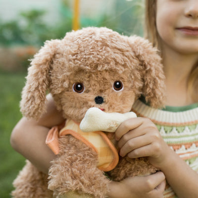 Soft golden plush Dinkum Dog with floppy ears holds a cream bone treat while being cuddled by a smiling child outdoors