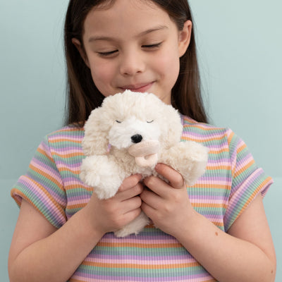 Young girl cuddling Belle a soft cream plush puppy with shaggy fur and a pink nose from Dinkum Pets against a pale blue background