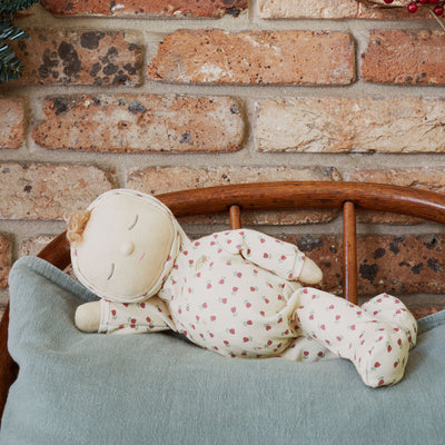 Sleeping Dozy Dinkum doll in a cream strawberry print outfit resting on a blue cushion in a wooden chair against a rustic brick wall by Dinkum Dolls by Olli Ella
