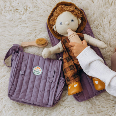 Dinkum Dolls fabric doll in a checked outfit beside a lavender changing bag and play mat with a toy brush on a soft rug