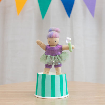 Holdie Circus acrobat doll in a purple tutu balancing on a green striped platform while holding a white and green circus prop beneath colorful bunting