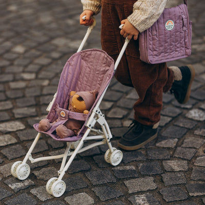 Child pushing a lavender quilted doll stroller with two soft toys inside on a cobblestone street from Dinkum Dolls by Olli Ella