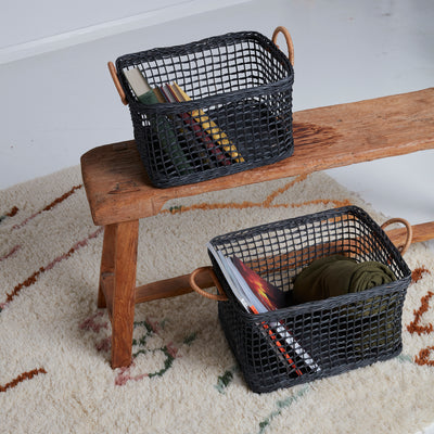 Two black rattan storage baskets with wrapped handles holding books and clothing on a wooden bench in a cosy home setting by Olli Ella Home