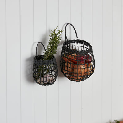 Olli Ella Home Ink rattan hanging basket duo displayed on a wall with onions in one basket and a leafy plant in the other