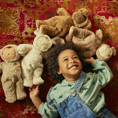 A smiling child lies on a patterned rug surrounded by soft plush dolls including a cream lamb toy from Cozy Dinkums for imaginative play