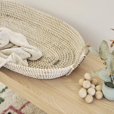 Natural seagrass changing basket with white woven detailing and a soft knitted blanket on a wooden table beside dried foliage and decorative wooden beads