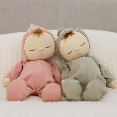 Two Dozy Dinkums dolls in pink and sage green hooded bodysuits sit together with sleepy embroidered faces on a soft white bed