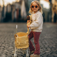 Child wearing orange sunglasses pushes a honey yellow doll changing bag in a pram while holding a teddy bear during imaginative play with Dinkum Dolls