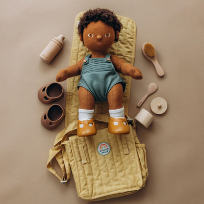 Dinkum Dolls doll in blue romper and yellow boots lying on a honey quilted changing mat with bottle shoes brush and changing accessories beside it