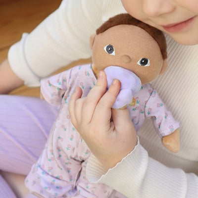 Child cuddling Binky Dinky Pip by Dinkum Dolls by Olli Ella wearing a floral outfit and holding a soft purple pacifier near its face