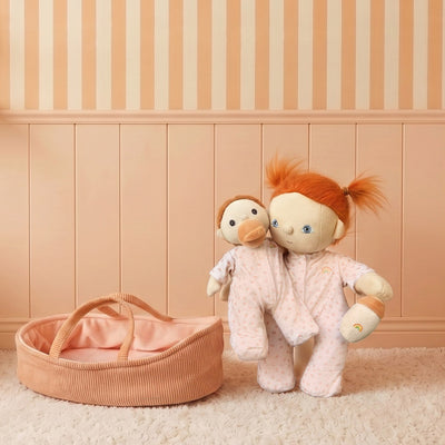 Dinkum Dolls by Olli Ella doll in soft pink floral pajamas cuddling a bunny toy beside a blush pink doll bassinet in a cosy nursery