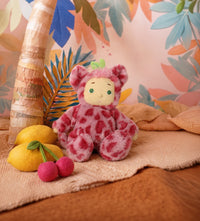 Pink spotted Cozy Dinkum plush doll with green eyes seated beside lemons and cherries in a tropical themed setting from Dinkum Dolls by Olli Ella