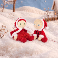 Two small Christmas-themed plush dolls sitting in a snowy outdoor scene. One doll wears a red dress and hood with white trim, and the other wears a red outfit with a Santa hat.