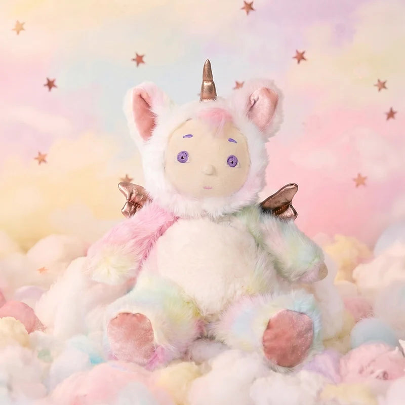 Cozy Dinkums plush unicorn doll with pastel rainbow fur and golden wings sitting among soft clouds against a dreamy pink starry backdrop