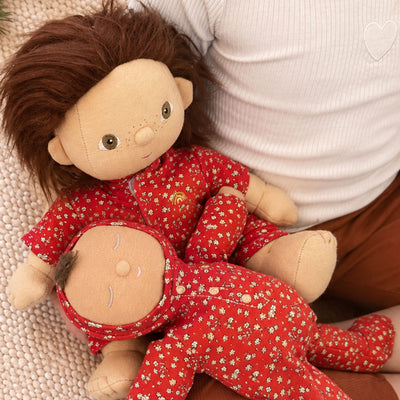 Two small plush dolls wearing matching red floral pajamas, posed on a person's lap. The dolls have beige faces and hands, one with brown hair and one with dark hair, against a white ribbed shirt and brown pants.