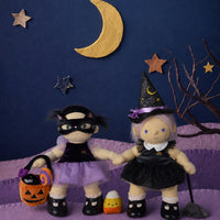 Two Halloween-themed dolls posed on a purple textured surface against a dark blue backdrop with a gold crescent moon, stars, and a bare tree branch. One doll wears a black costume with a purple skirt and holds an orange jack-o'-lantern bucket.