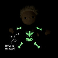 A dark plush doll with short blonde hair wearing a black outfit that glows green with a skeleton bone and rib pattern, shown on a black background with the text 'GLOWS IN THE DARK!'.