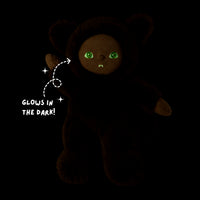 A dark plush teddy bear with glowing green eyes and a small stitched smile, shown on a black background with the text 'GLOWS IN THE DARK!' and sparkles pointing to its eyes.