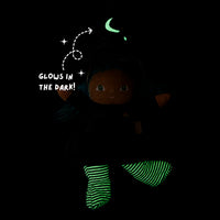 A dark plush doll wearing a black outfit and glowing green striped socks, with a glowing green crescent moon and stars on the hat. White text reads 'GLOWS IN THE DARK!' on a black background.