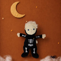 A Halloween plush doll dressed in a black skeleton jumpsuit with white bone details, posed against a textured brown background with a yellow crescent moon, stars, fake white cobwebs, and a small black spider decoration.
