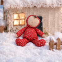 Soft baby doll with sleepy embroidered closed eyes, a smiling mouth and a tuft of dark brown hair, wearing a red footed sleepsuit and matching bonnet with a ditsy floral print, button placket, and a small embroidered rainbow.