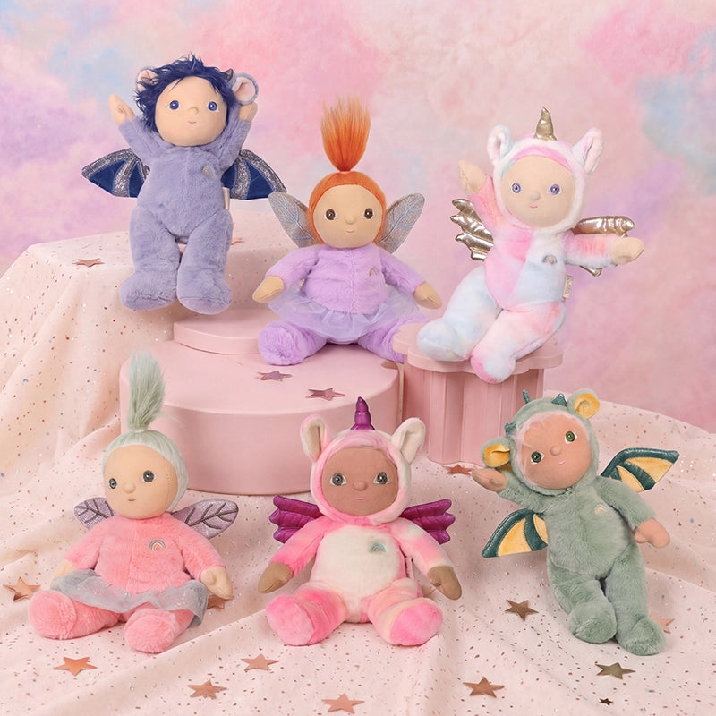 Six Dinky Dinkums soft dolls in colorful fantasy costumes including unicorns and fairies arranged on pink pedestals against a dreamy pastel backdrop