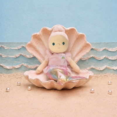 A doll with a pink hat and pink dress sits inside a large pink clam shell on a sandy beach set with a blue textured background and small pearls.