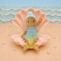 A mermaid doll with a blue and gold tail sits inside a large pink clam shell on a sandy beach backdrop with blue ocean waves behind it.