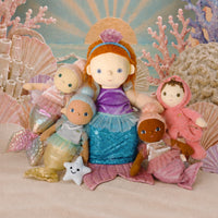 A group of mermaid-themed plush dolls arranged on a sandy surface with seashells, coral, and a painted ocean backdrop. The central doll has red hair, a purple top, and a blue-green mermaid tail, while the surrounding dolls wear pink, gold, and peach outfits.
