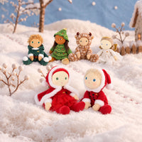 A set of soft plush dolls styled in a snowy winter scene. In the foreground, two dolls dressed in red Santa-style outfits sit facing each other.